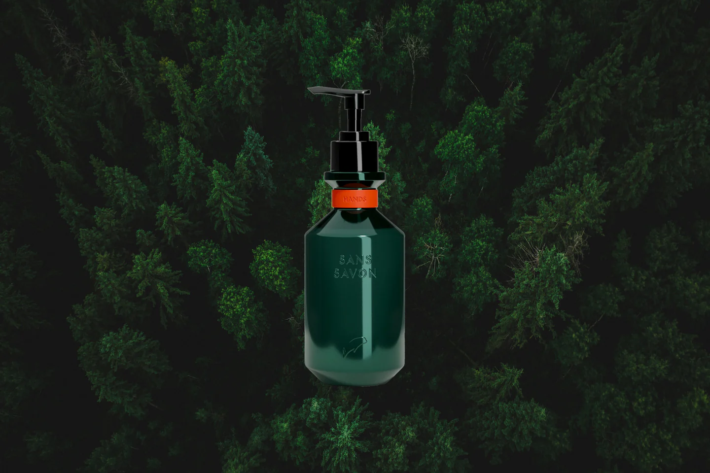 A dark green, cylindrical bottle of Sans Savon hand wash with a black pump dispenser is digitally placed against a blurred, overhead view of a dense evergreen forest. The bottle features a vibrant orange band around its neck with the word "HANDS" embossed on it. The brand name "SANS SAVON" is vertically printed on the front of the bottle. The blurred forest canopy in the background creates a textured, dark green backdrop, likely aiming to evoke a sense of natural purity and connect the product with the outdoors.