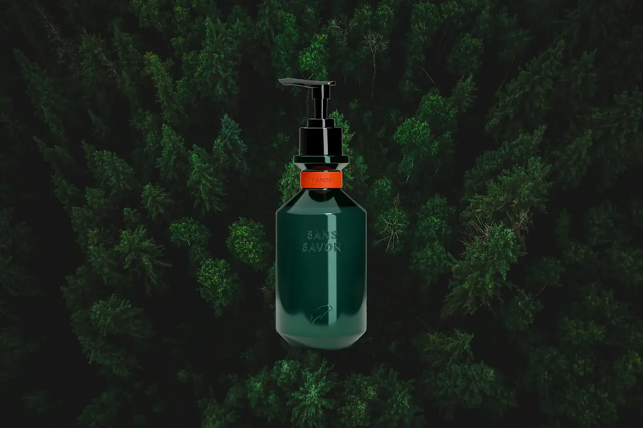 A dark green, cylindrical bottle of Sans Savon hand wash with a black pump dispenser is digitally placed against a blurred, overhead view of a dense evergreen forest. The bottle features a vibrant orange band around its neck with the word "HANDS" embossed on it. The brand name "SANS SAVON" is vertically printed on the front of the bottle. The blurred forest canopy in the background creates a textured, dark green backdrop, likely aiming to evoke a sense of natural purity and connect the product with the outdoors.