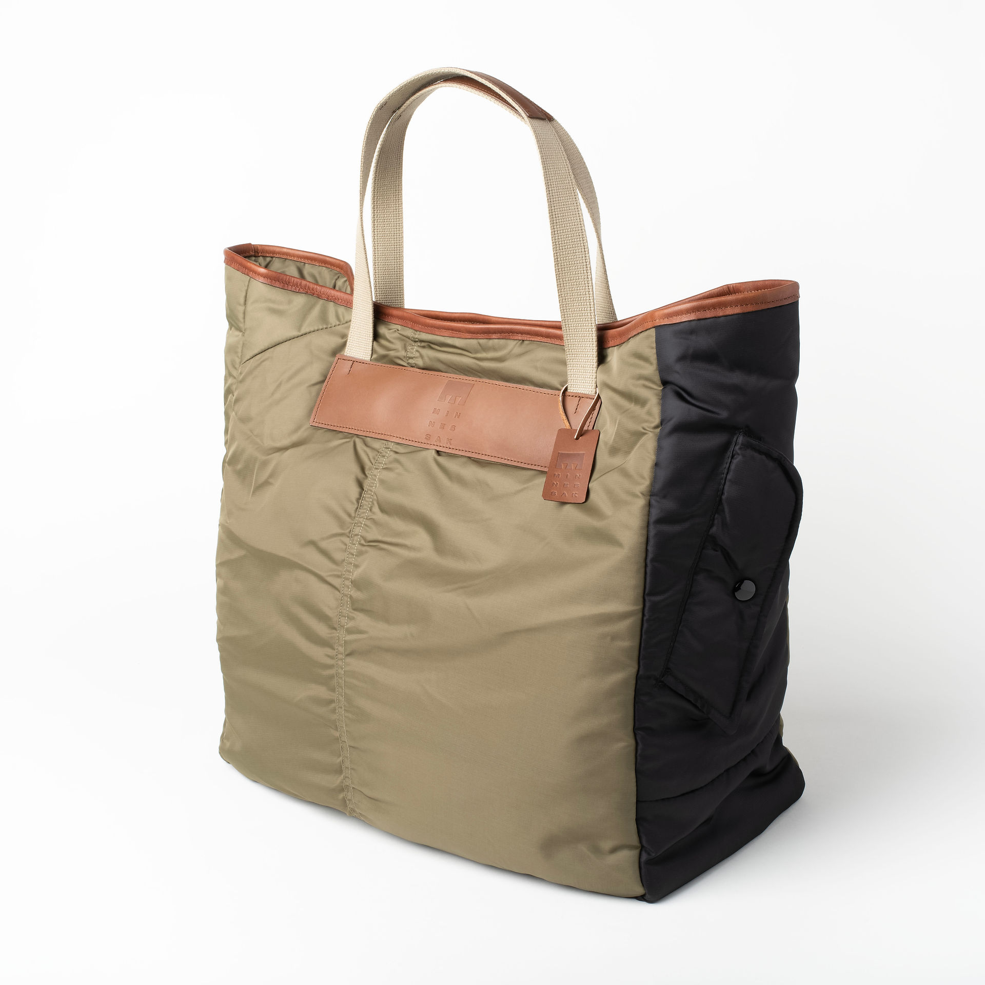 A Minnessak Tote Bag is presented against a light grey background. The tote features a combination of materials and colors. The main body is a light olive green, quilted fabric. One side panel is black and also quilted, featuring a large patch pocket with a flap and snap closure. The top edge of the bag is trimmed with brown leather. The tote has two cream-colored, wide nylon straps that extend from the top. A brown leather tag with embossed branding hangs from one of the straps. The lighting is soft and even, highlighting the textures and the different sections of the bag.
