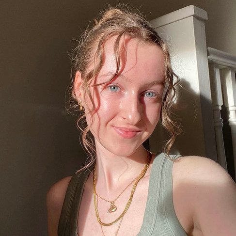 A sunlit selfie of Ella, a nail influencer, shows her smiling warmly at the camera. Her light brown, wavy hair is partially pulled back, with loose curls framing her face. She has fair skin and bright blue eyes. She is wearing a ribbed olive-green tank top and layered gold necklaces, one with a small key pendant and another with an elephant charm. The background is a softly lit interior space with a white staircase railing visible on the right. The sunlight creates a warm glow on her face and hair.