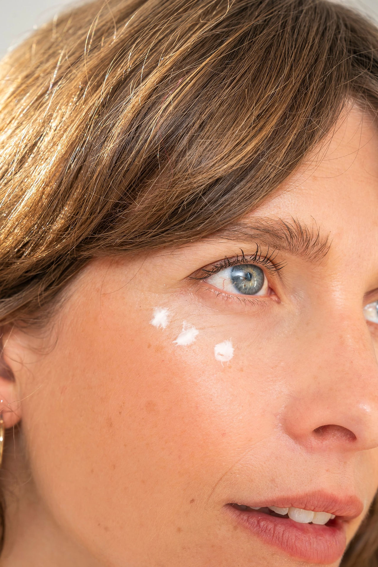 A close-up shot focuses on the side profile of a fair-skinned woman's face, highlighting her eye area. She has light blue eyes, visible eyelashes, and light brown hair with subtle highlights. Three small dots of white cream or lotion are applied beneath her left eye, near the cheekbone. The lighting is bright, casting soft shadows and emphasizing the texture of her skin. A hint of a gold hoop earring is visible near her ear. Her expression is neutral, and she appears to be looking slightly upwards and to the right.
