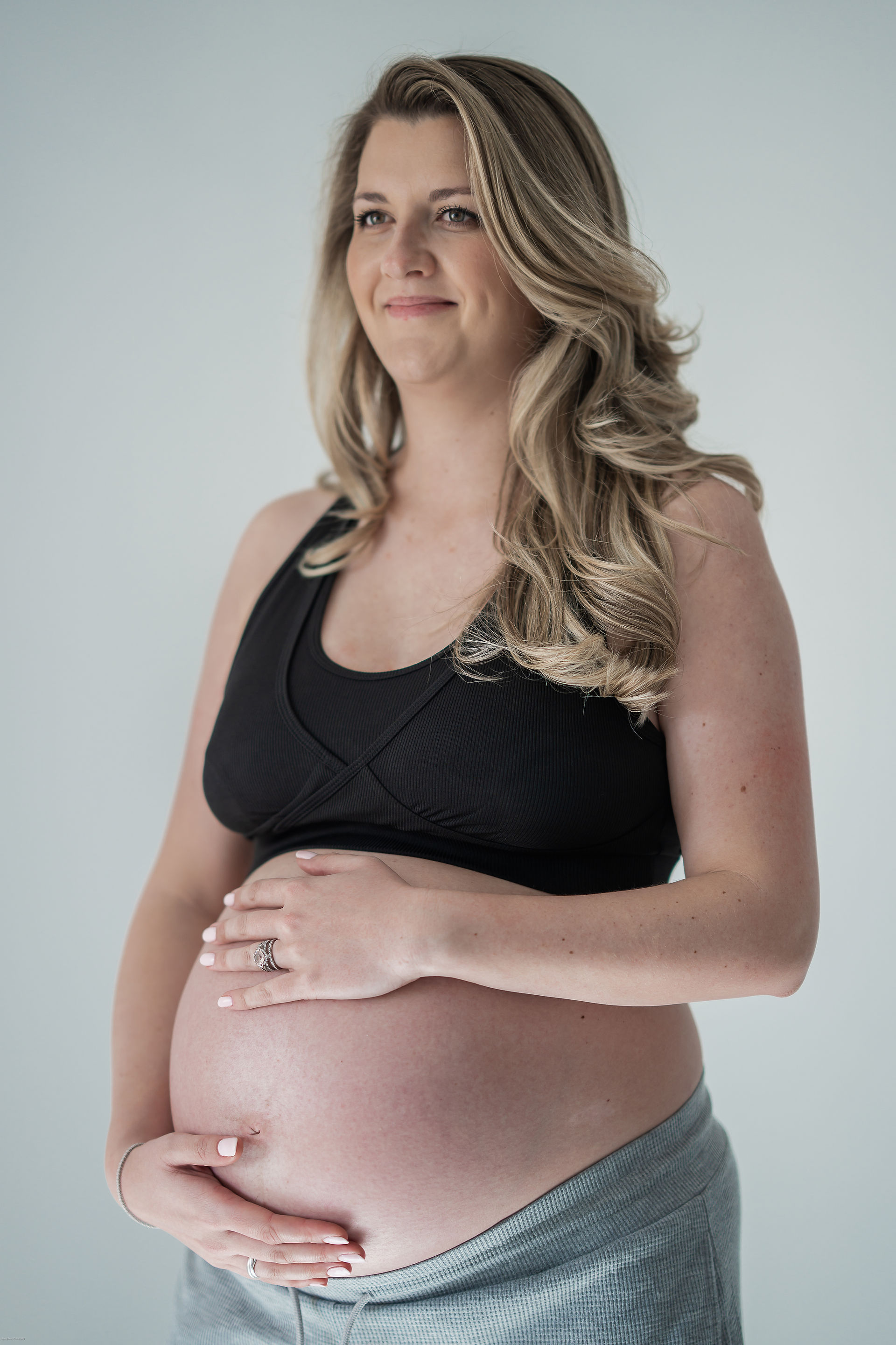 A pregnant woman with long, wavy blonde hair is shown from the chest up against a soft grey background. She is wearing a black tank top and grey sweatpants pulled down to reveal her prominent baby bump. Her hands gently cradle her belly, and she is looking off to her left with a serene smile. She is wearing a ring on her left ring finger and a bracelet on her left wrist. The lighting is soft and even, highlighting her peaceful expression and the curves of her pregnant form.