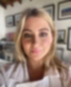 A close-up portrait of Lark with blonde, shoulder-length hair and light-colored eyes. She has a natural makeup look, with rosy cheeks and glossy lips. She is wearing a white collared shirt. The background shows a room with framed pictures on the wall and a shelf with records or books.