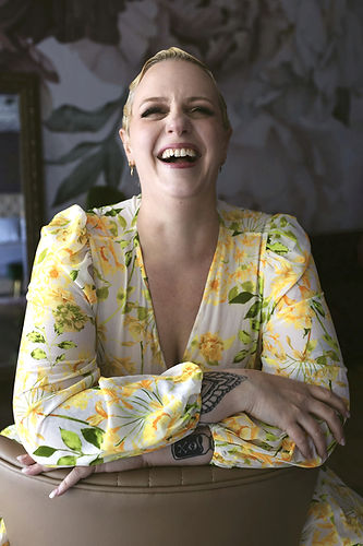 Richelle in a yellow floral dress, with her arms crossed and leaning to the left. She is laughing a toothy laugh. Her hair is blonde and styled in finger waves.