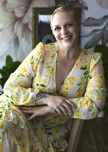 Richelle in a yellow floral dress, with her arms crossed and leaning to the left. She has a smile on her face. Her hair is blonde and styled in finger waves.