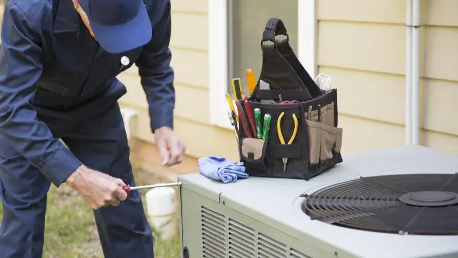 HVAC tech using a screwdriver on a sysem outside. He has his tool bag sitting on top of the system.