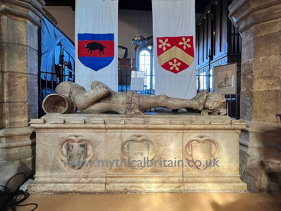 Effigy of a fourteeth century knight on a tomb chest featuring three shields depicting heraldic devices. Above the knight can be seen two modern decorative banners while at the knight's feet can be seen a cheerful carving of a lion.