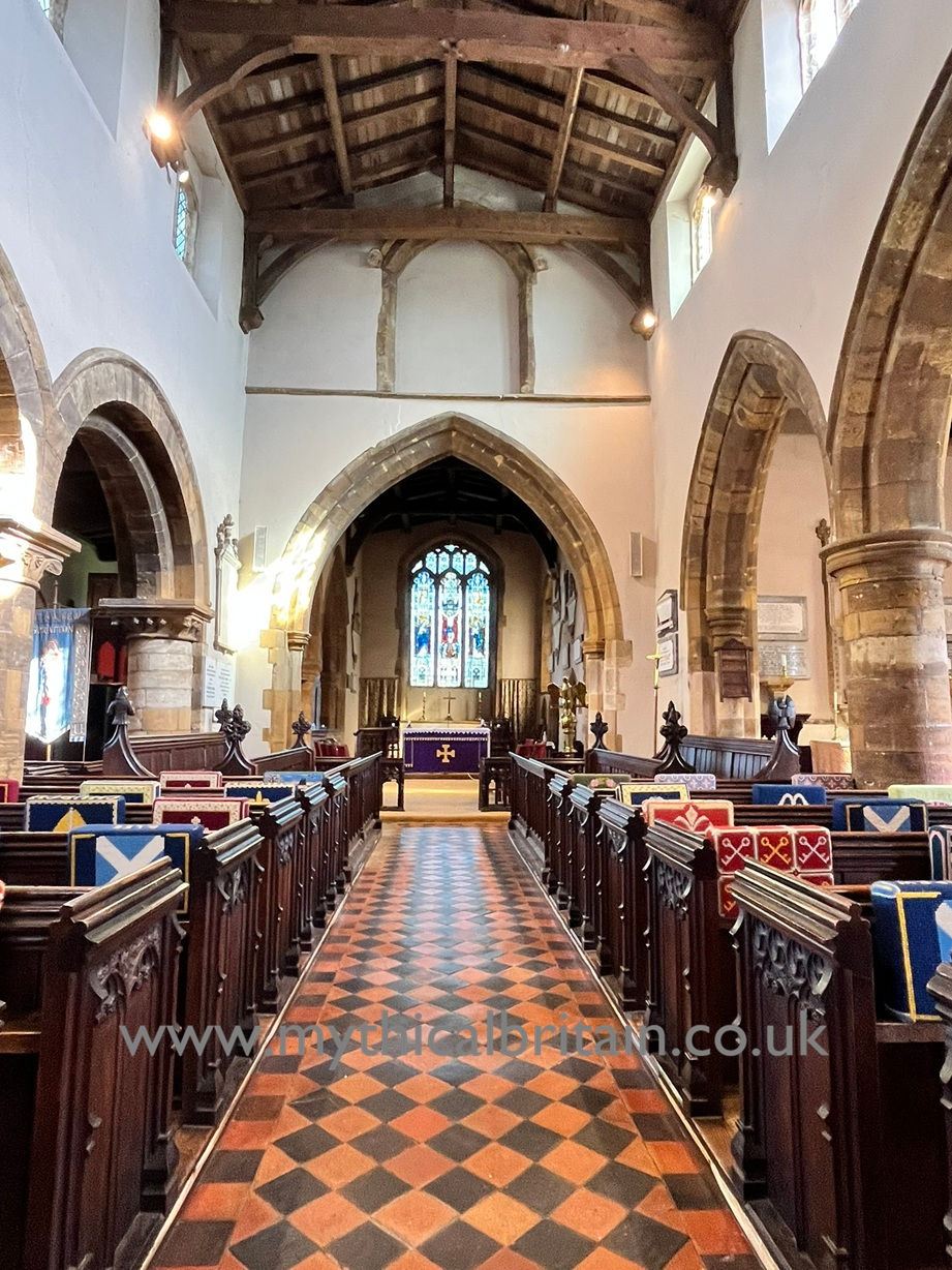 Interior of church showing pews, tiled floor, pillars and arches and view towards the chancel with a stained glass window in the distance and a niche above the chancel arch.
