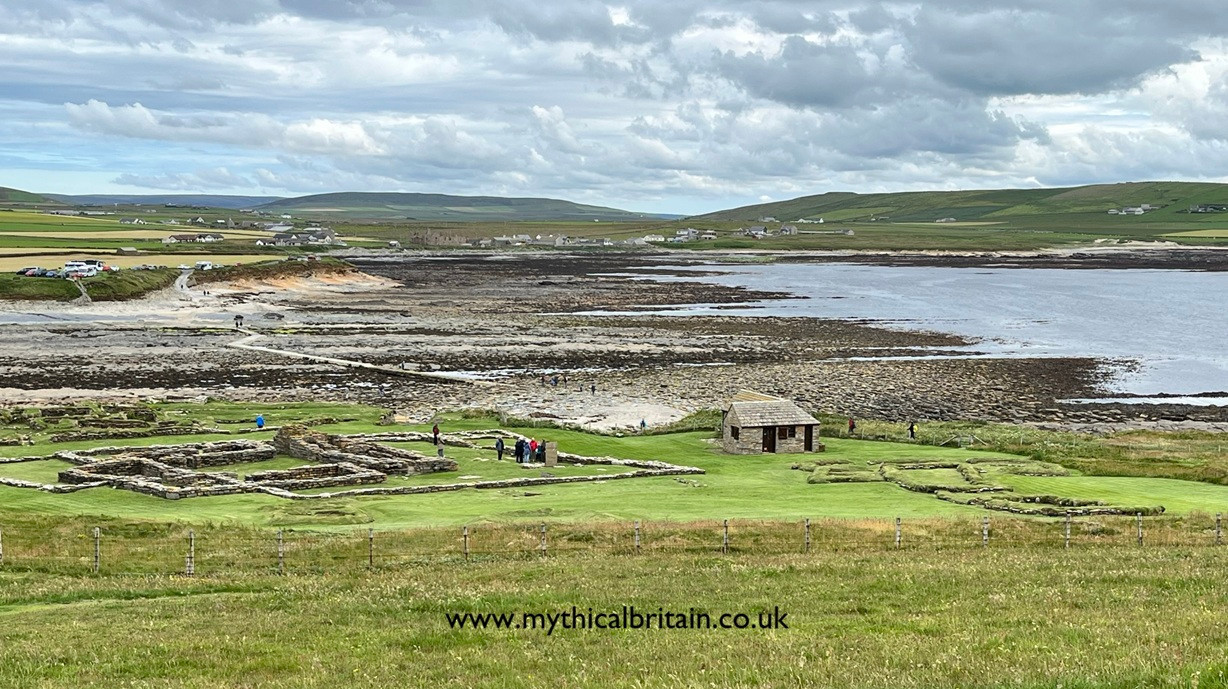 View over some ruins towards a causeway crossing a stony beach towards a distant car park.