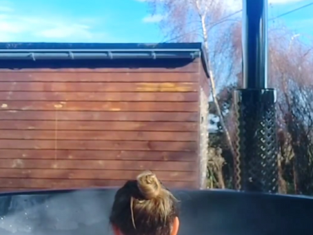 Jeune femme vue de dos, avec un chignon, se prélassant dans les bulle d'un spa ou bian nordique en extérieur, avec une tiny house en bois et le ciel bleu en arrière-plan