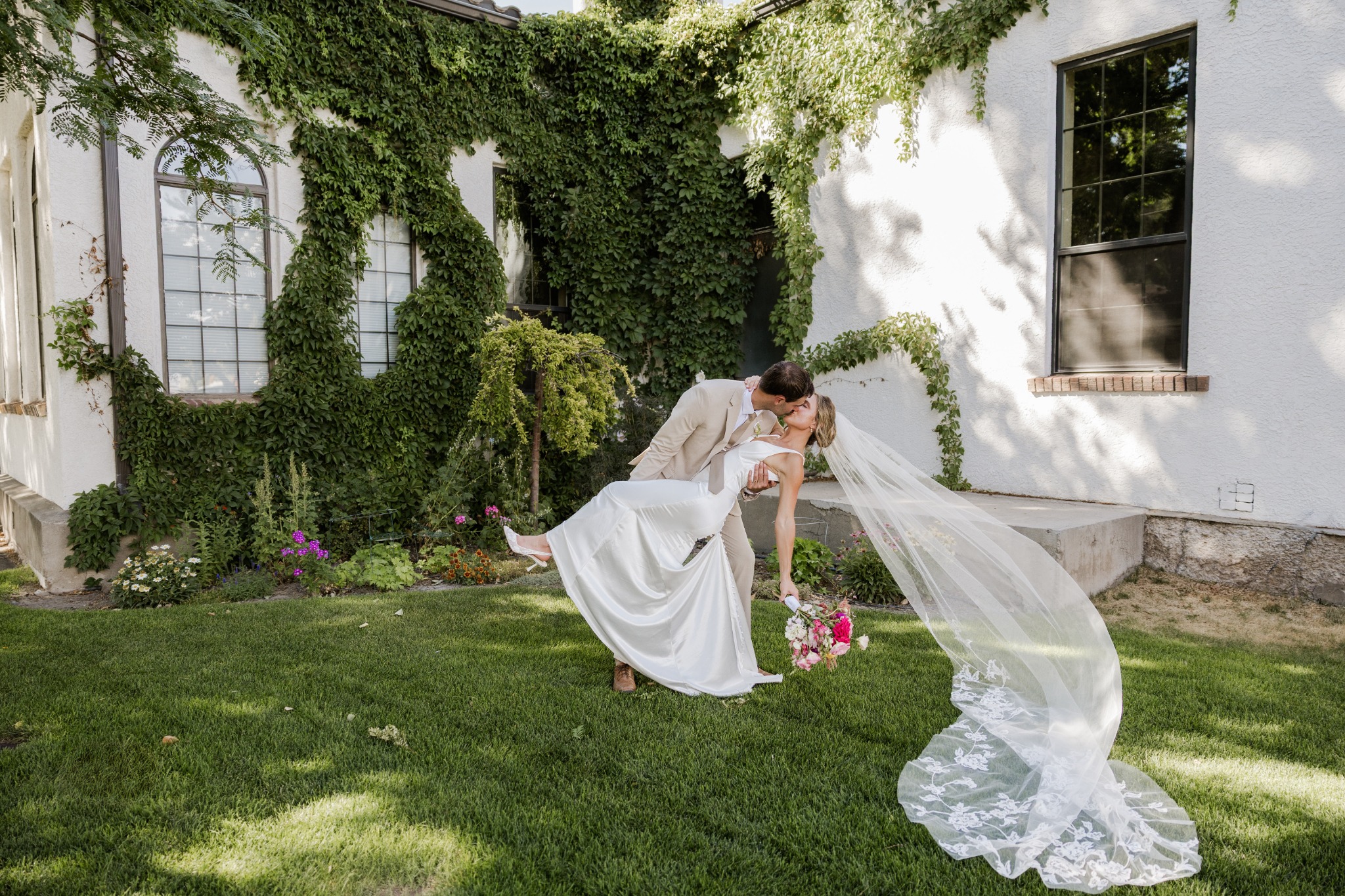 A bride and groom leaning into a kiss beneath a flowing veil in an ivy-lined garden