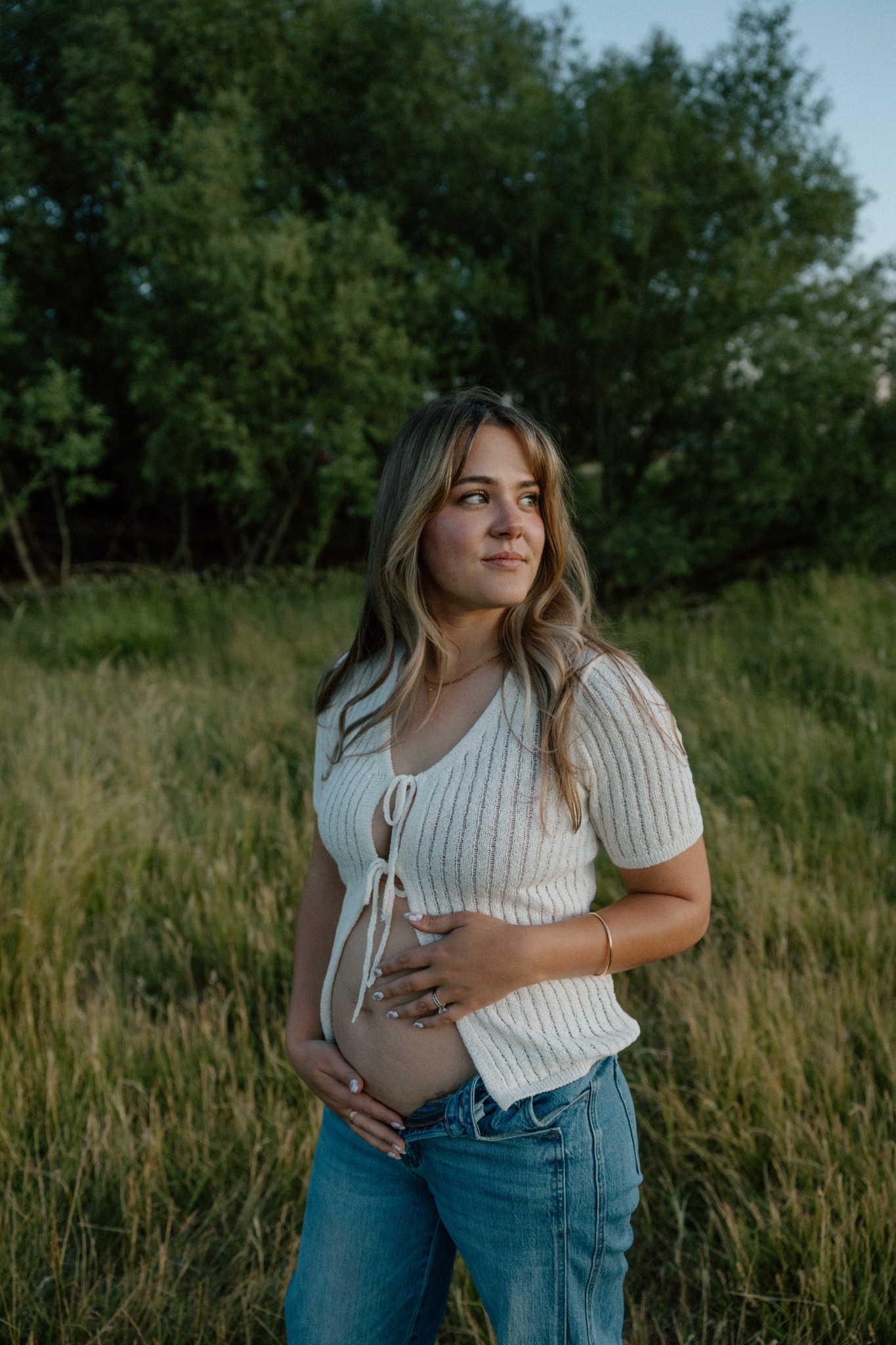 Julia resting her hands on her baby bump in a grassy field beside trees