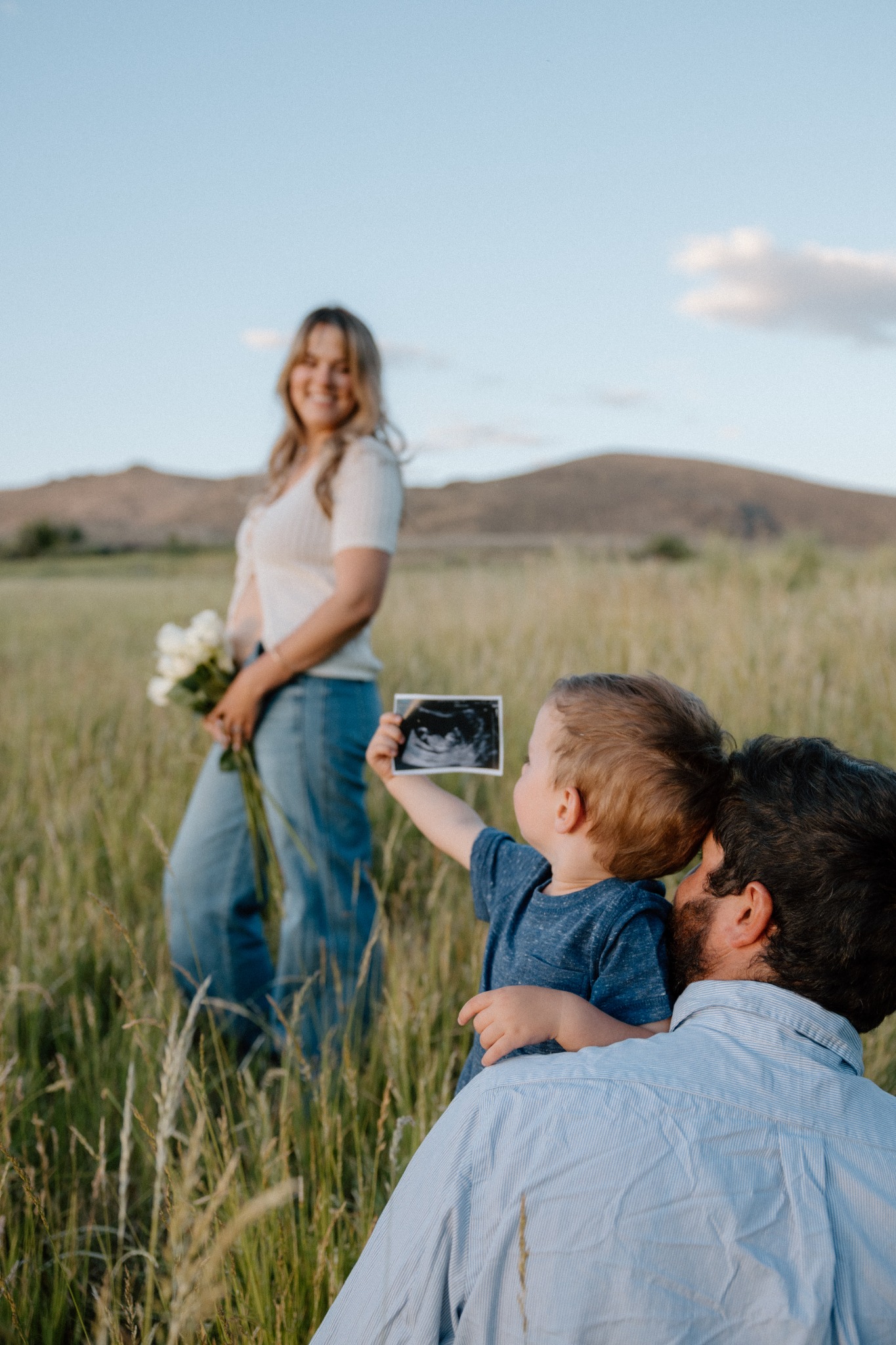 A child holding an ultrasound photograph beside a parent, with Julia in the meadow behind them