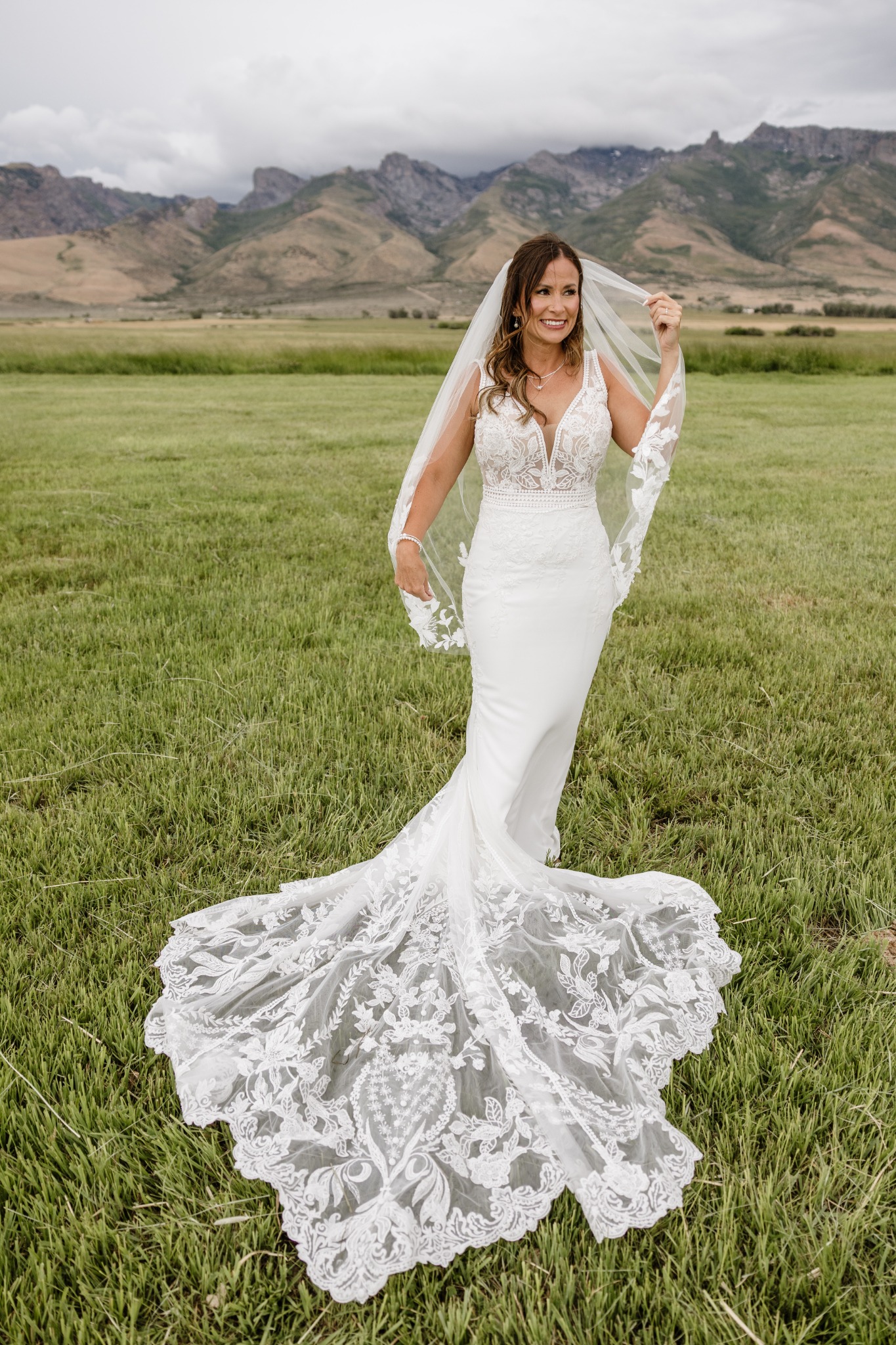 Amy lifting her veil in a meadow, with her lace train and mountains in view