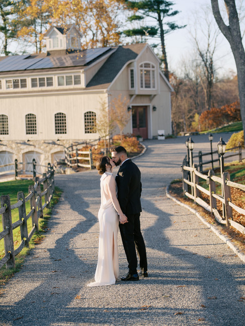 Bride and groom kissing on a tree-lined driveway in front of a barn at a countryside wedding venue
