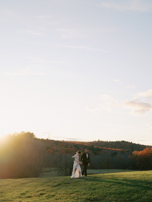 Bride and groom walking across a sunlit meadow at sunset during a fall wedding with rolling hills in the background