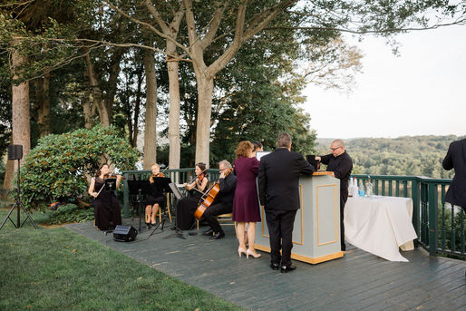 saint clements castle cocktail hour on the Waterford side overlooking the connecticut river. in the foreground, a male bartender is serving guests. in the background, an amplified string quartet is performing in all black. 