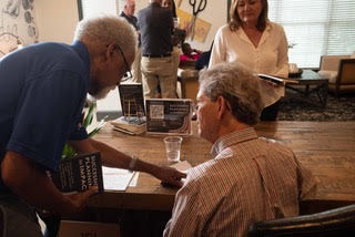 Chairman leans over a table talking to Harry T Jones; a woman stands behind. Open laptop, papers, and a calm indoor meeting setting.
