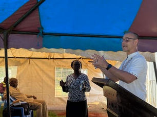 Man speaking at outdoor ministry event under colorful tent with audience listening in Kenya