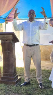 Pastor speaking outdoors under canopy with raised hands gesturing during teaching session on Biblical truth
