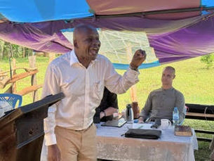 Man in white shirt speaking at outdoor event under colorful canopy with people seated at table behind him on grassy field
