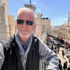 David Oaks with gray hair and beard wearing sunglasses standing on balcony overlooking busy marketplace with stone buildings