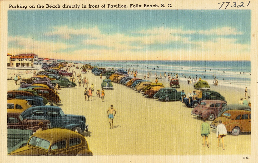 Retro cars parked on a crowded beach, people walking and relaxing by the sea under a clear sky, with pavilion buildings in the distance.