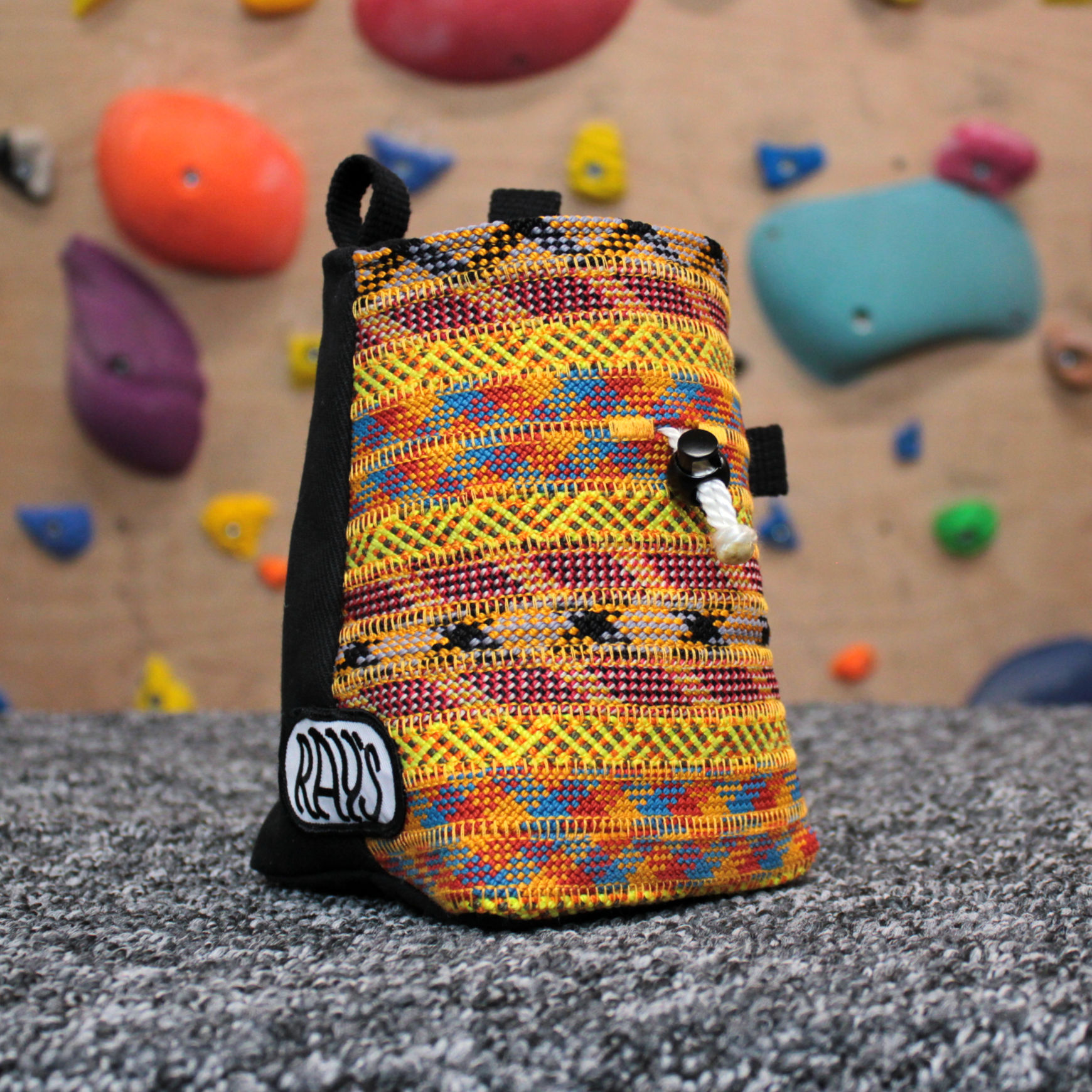 A chalk bag in warm tones is sat in front of a climbing wall, it is handmade by Ray's Climbing from repurposed climbing ropes