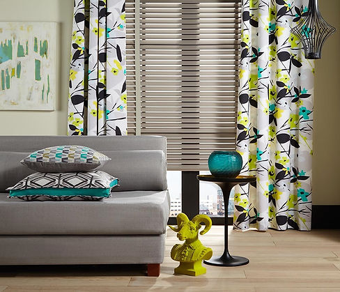 A grey sofa, with 2 scatter cushions on it, in front of a big window with wood blind and floor length curtains