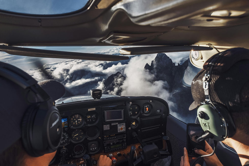 Two pilots fly a small plane through clouds and over mountains. The cockpit displays instruments, with blue sky visible through windows.