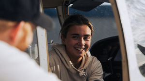 Woman smiling inside a small plane cockpit, wearing a beige hoodie. Man in a cap stands outside, daytime with blue sky in background.