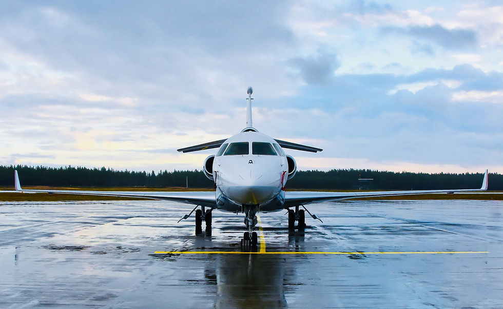 Front view of a white jet on a wet runway under a cloudy sky. The background shows a distant treeline and a hint of blue sky.