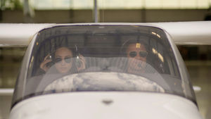 Two people in aviator sunglasses sit in a small aircraft cockpit. They prepare for flight, adjusting headsets, with hangar visible outside.