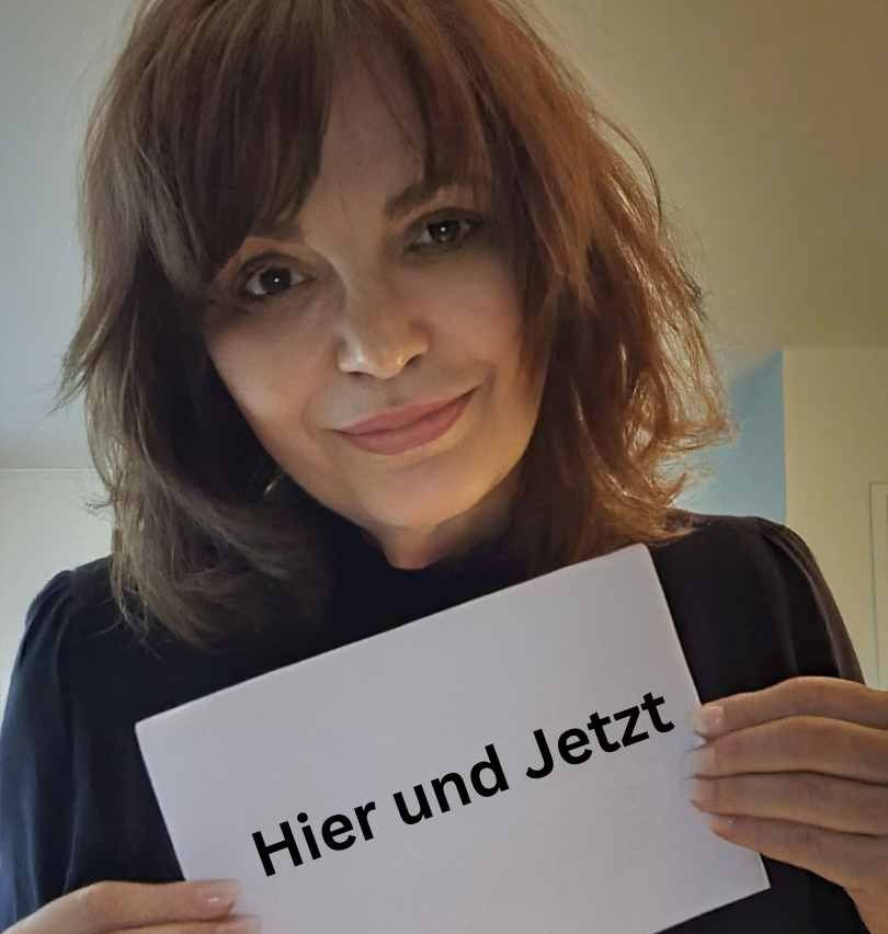 Eine Frau hält ein Schild mit der Aufschrift "Hier und Jetzt" in die Kamera