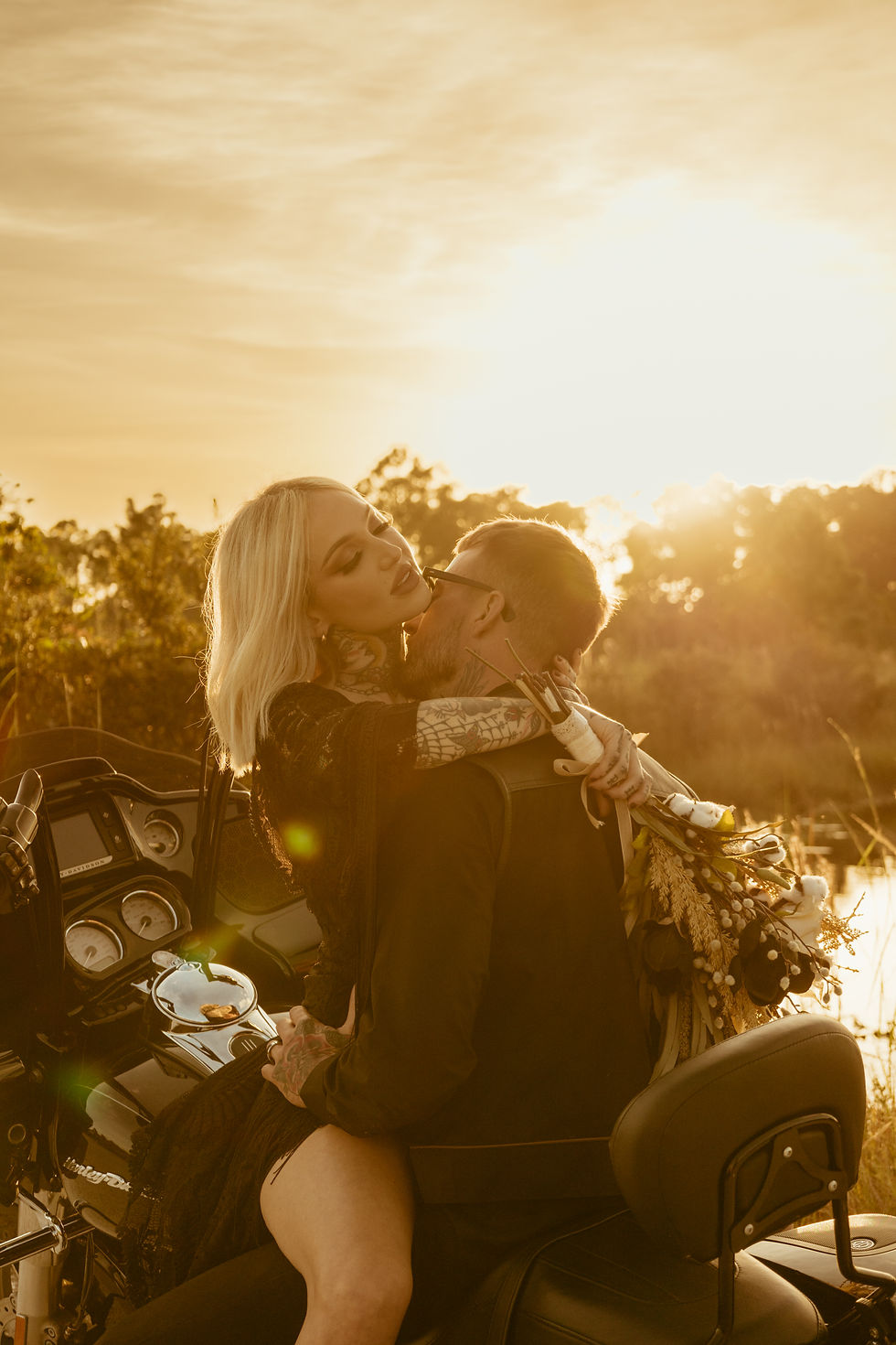 A couple embraces on a motorcycle at sunset, surrounded by nature. Warm golden tones, with a tattooed hand holding a bouquet. Romantic mood.