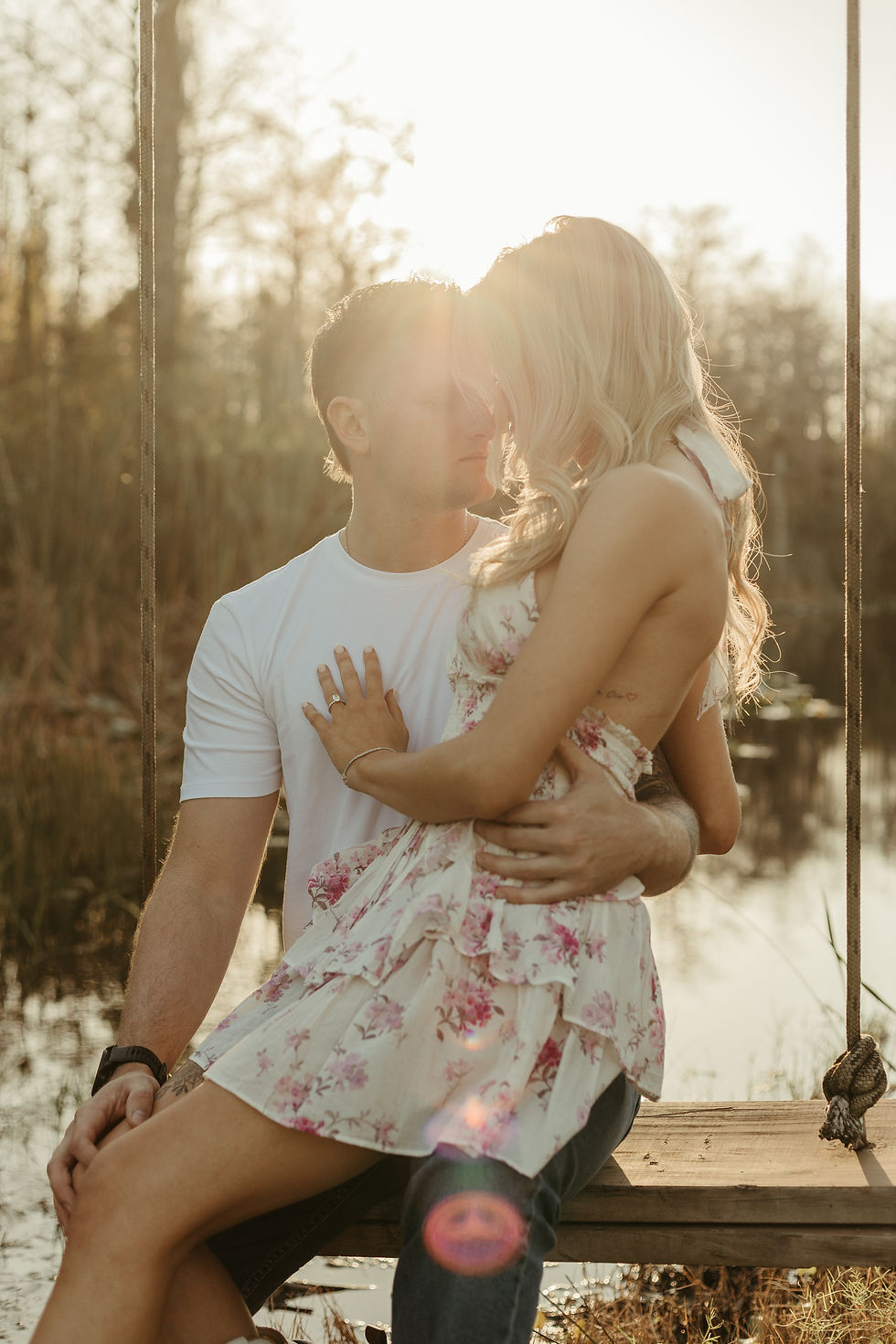 Couple embracing on a swing by a lake at sunset, the woman in a floral dress. Sunlight creates a warm, romantic atmosphere.