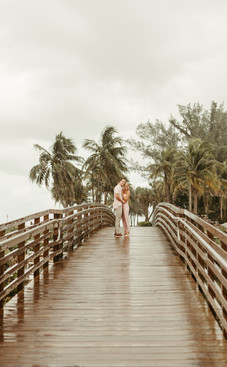 A couple holding hands walks on a wet wooden boardwalk in rainy weather. Palm trees in the background, both smiling, wearing pink attire.