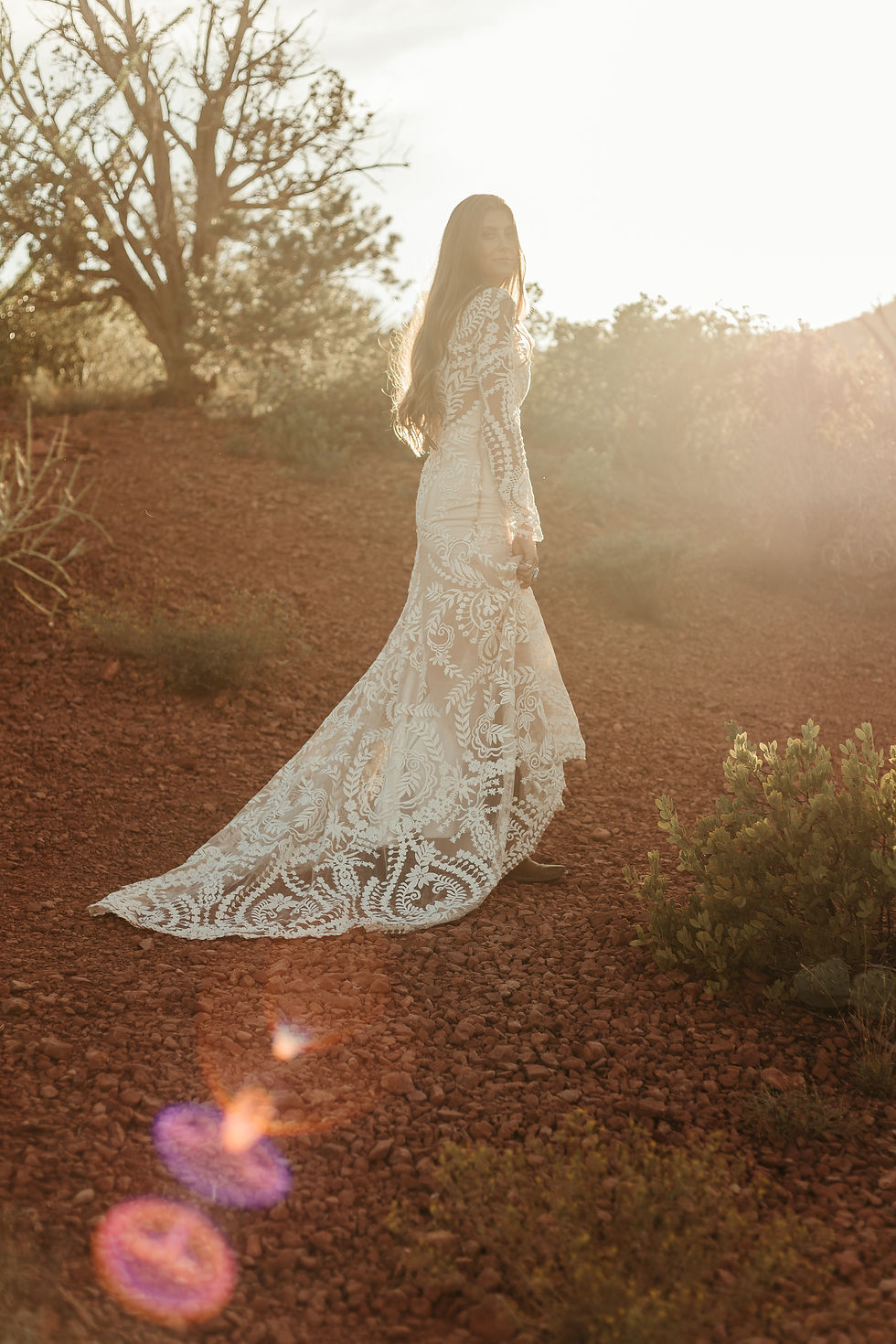 Woman in a white lace dress walks on a sunlit red dirt path, surrounded by greenery and trees, with a warm, dreamy atmosphere.