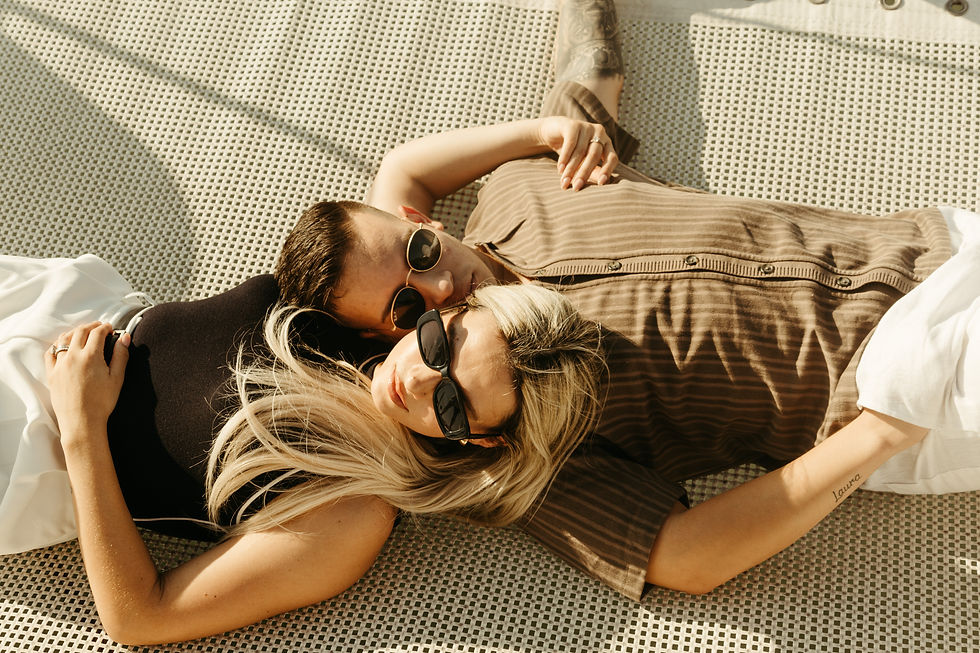 Two people wearing sunglasses lie on a patterned surface, relaxed and cozy in the sunlight. One has a visible tattoo. Light and relaxed mood.