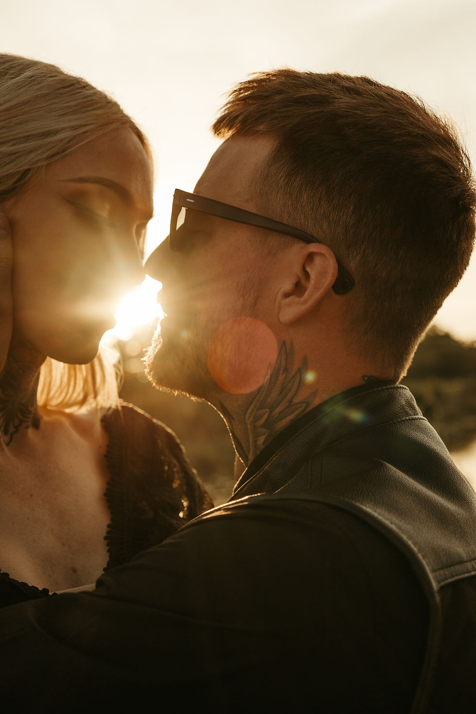 Close-up of a couple about to kiss at sunset, sun flare between them. The man wears sunglasses and a leather jacket, the scene feels romantic.