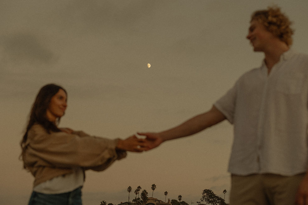 A couple holds hands under a dusk sky with a visible moon. Background shows palm trees and a distant house for a serene mood.