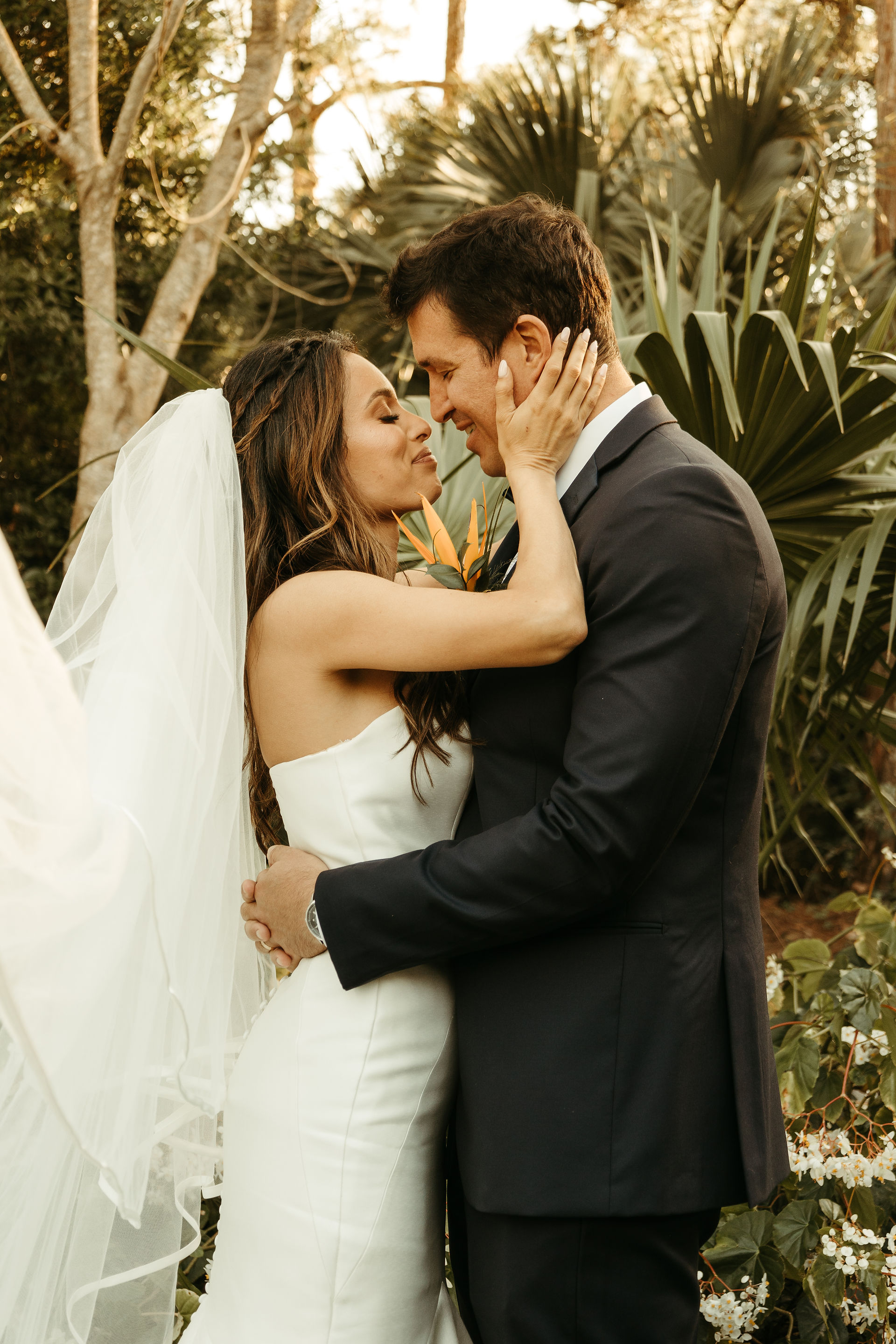 Bride and groom smiling, holding tropical bouquet in garden with white statue and palm trees. Bright sunny day, elegant white dress.