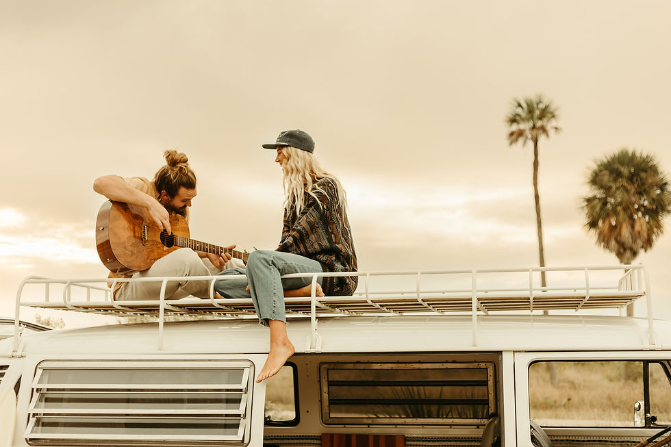 Man playing guitar and woman sitting on a van roof at sunset. Desert setting with palm trees, relaxed mood, earthy colors.