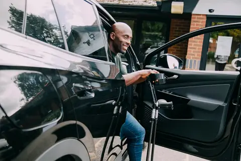 Smiling Motability customer using crutches while getting out of a parked car outside a building