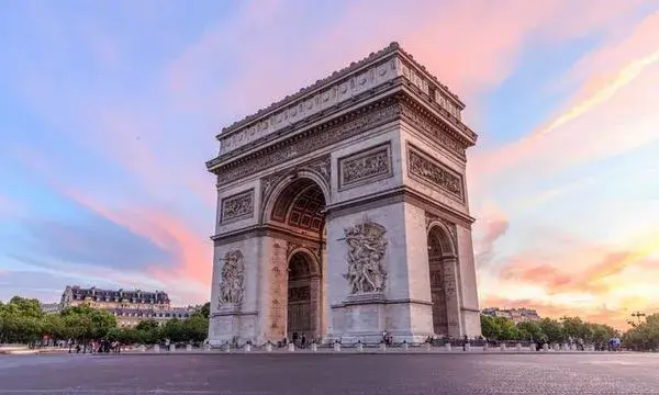 The Arc de Triomphe stands majestically at sunset, framed by pink and blue skies. Surrounding trees add a touch of nature to the historic scene.