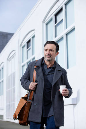 John Cassini walks down the street with a brown shoulder bag and white coffee cup in hand
