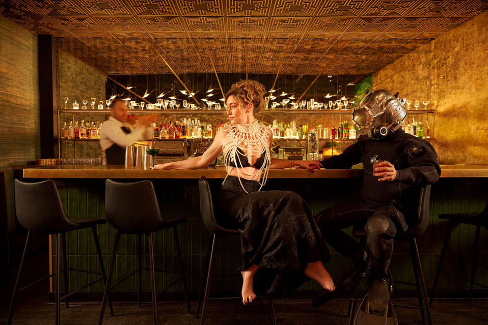 Camila Montgomery sits on a barstool with her arms up on the bar as she looks to left of frame while Rohano dressed in deep sea diving gear looks at her