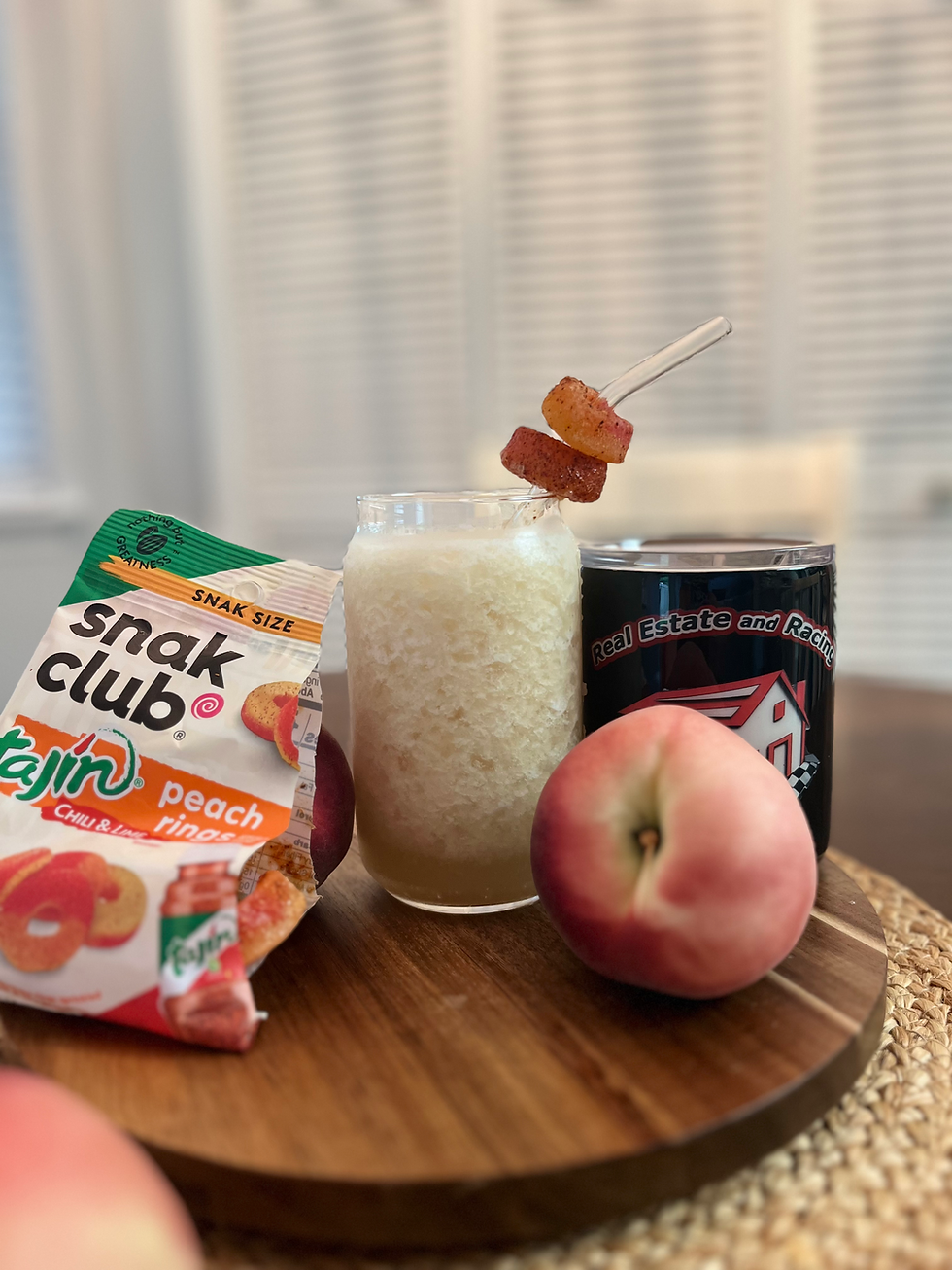 Peach drink with a skewer of peach rings on a table, surrounded by fresh peaches, a "Snak Club" bag, and a cup. Cozy indoor setting.