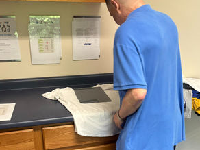 A man in a blue shirt using a black shirt-folder board to fold a white shirt on a navy countertop