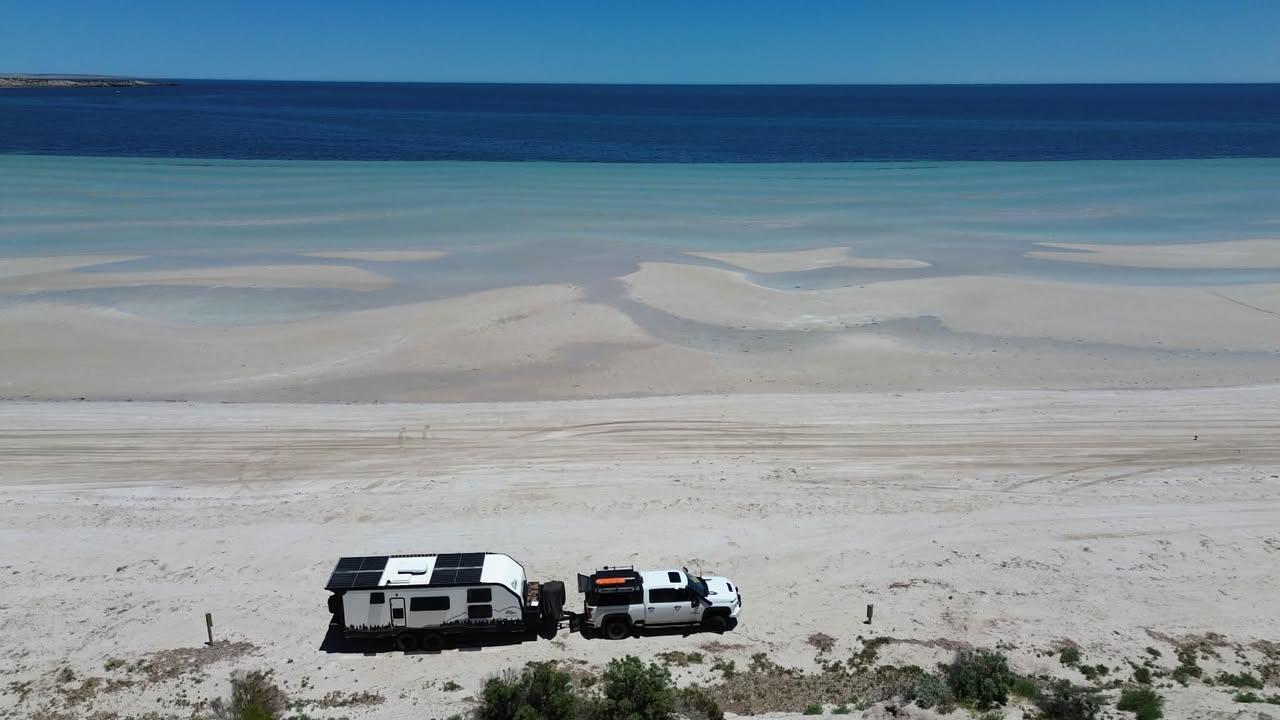 An Offroad Grit RV and ute on a white-sand beach by turquoise water, from the air